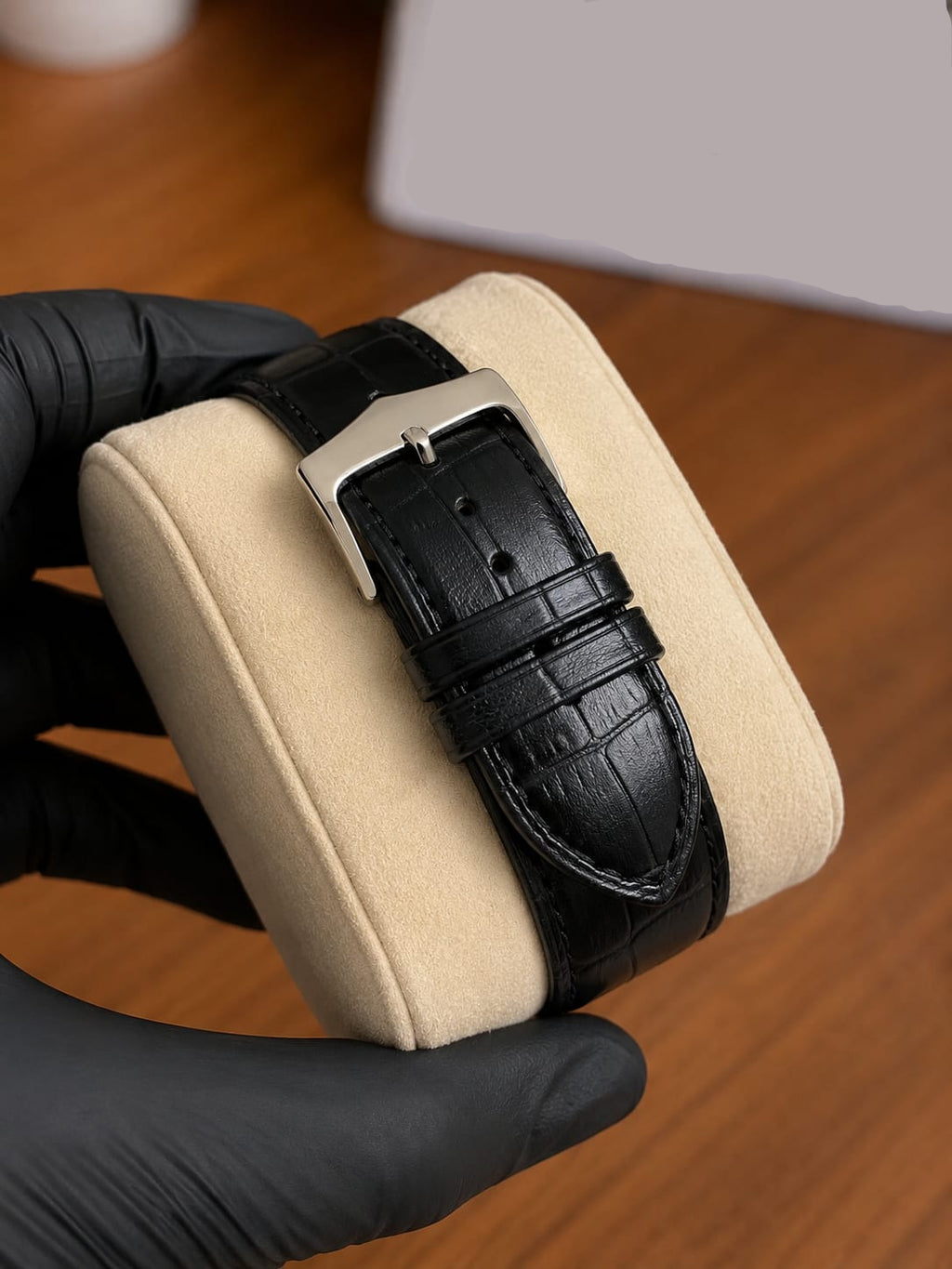 A close-up of a black leather watch strap with a silver buckle, held by a hand wearing a black glove over a cream-colored cushion on a wooden table.